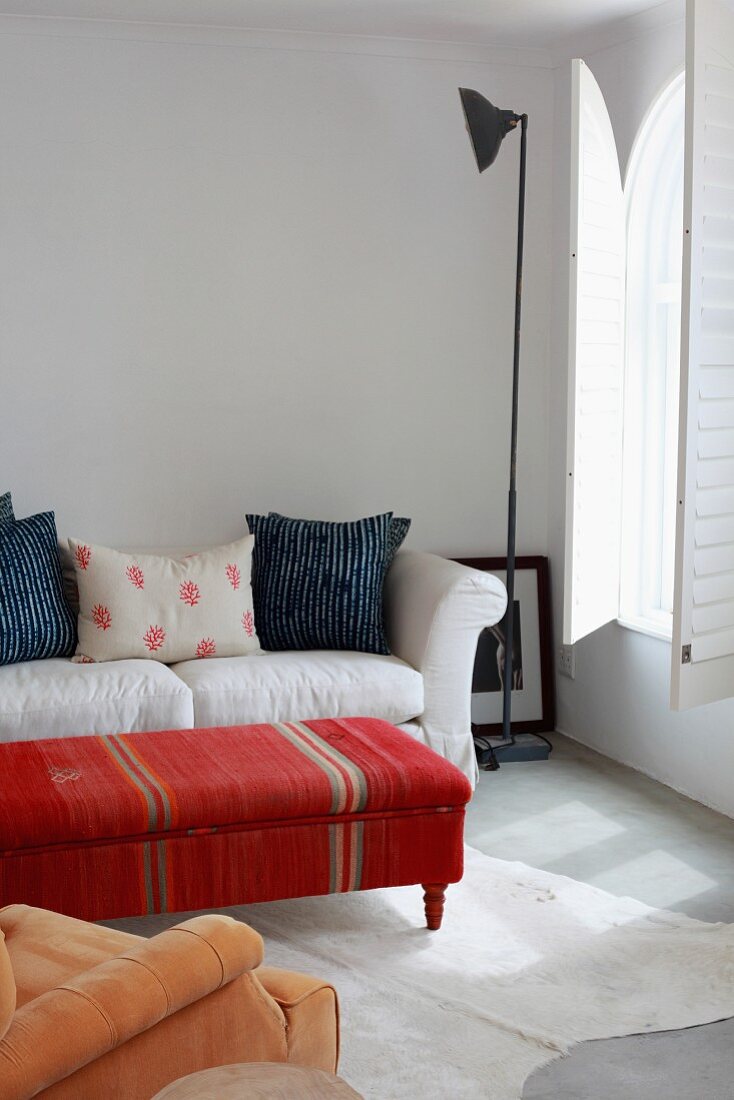 Couchtisch mit folkloristischem Bezug vor hellem Sofa und Retro Stehleuchte in minimalistischem Zimmer mit Innenläden an Rundbogenfenster