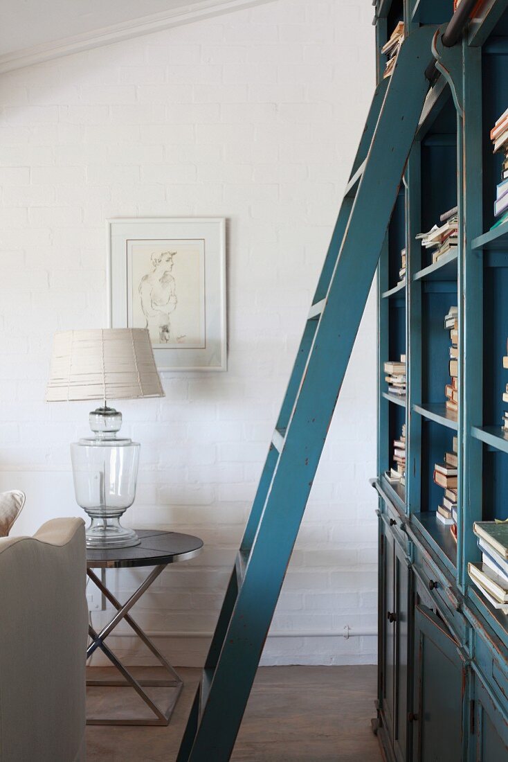 Blue, lacquer ladder in front of a bookcase and table lamp with earth tone lamp shade on a modern side table by the wall