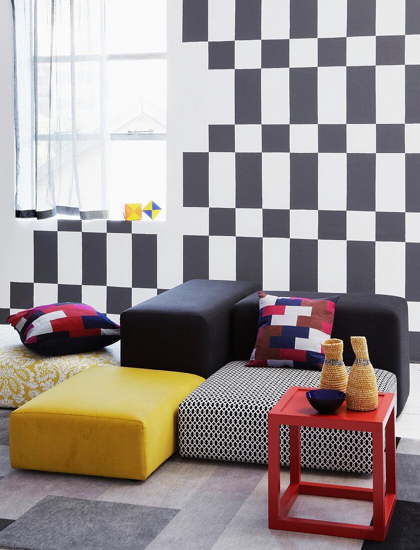 Red side table and floor cushions in a variety of colors in front of a wall with a black and white, geometric pattern