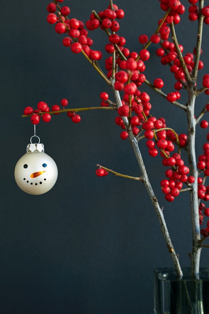 Weihnachtskugel mit aufgemaltem Schneemanngesicht an dekorativem Stechpalmenzweig