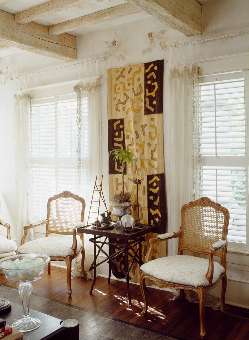 Side table in front of ethnic wall hanging and antique chairs in front of windows with louver blinds
