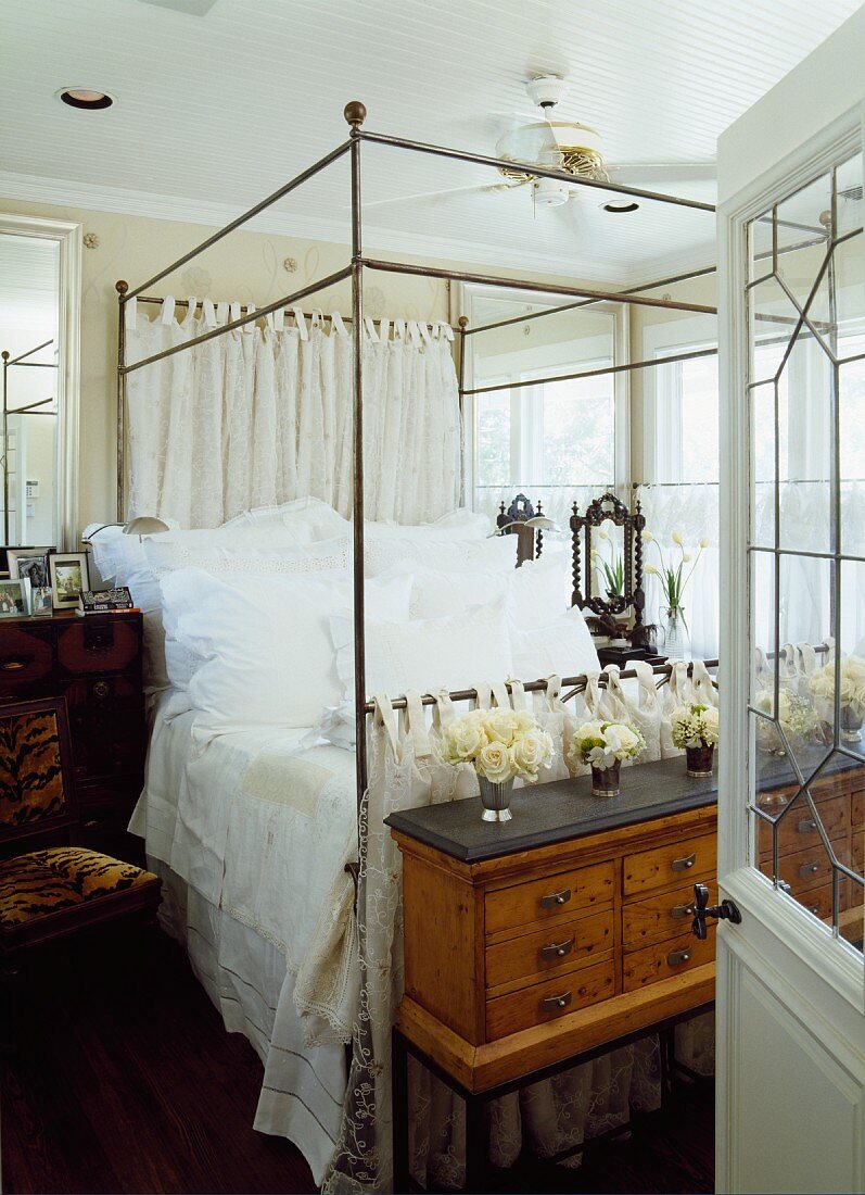 Blick durch offene Tür auf Himmelbett mit Metallgestell; an Bettende antike Kommode mit Blumensträusschen