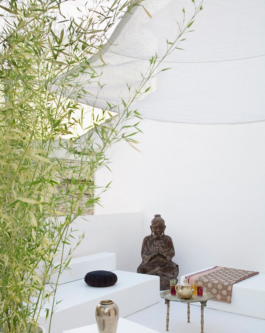 Seating area as continuous, white spatial sculpture half hidden behind bamboo with figure of Buddha and small Oriental tea table