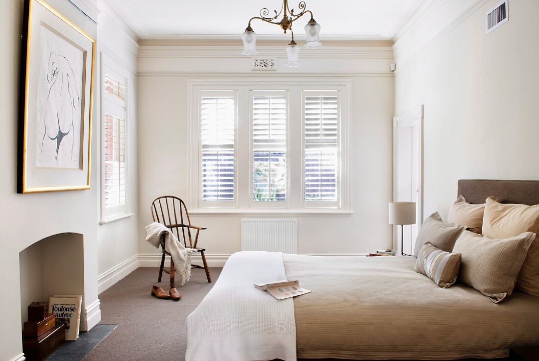 Pale bedroom with stucco friezes and exterior louver blinds; bed with upholstered headboard and bed linen in shades of beige and clothing on Shaker-style chair
