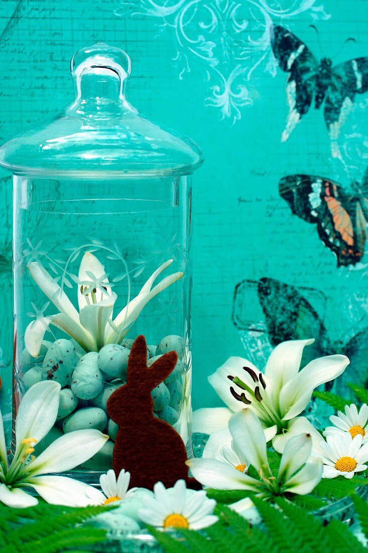 Dekoration mit Filzhase vor Glasgefäss; Lilienblüten und Margeriten vor blauem Hintergrund mit Schmetterling