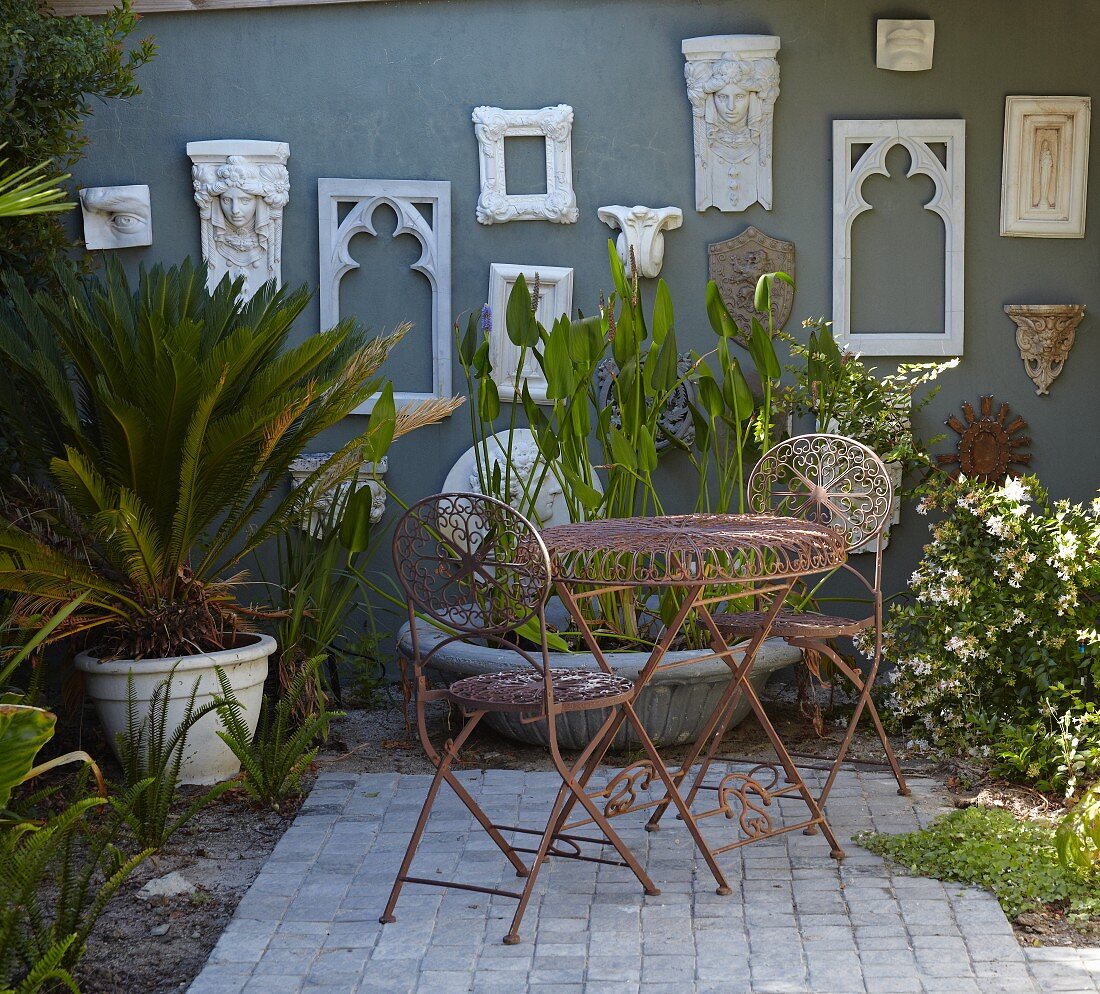 Delicate, metal outdoor furniture and potted plants on terrace in front of stylised antique frames on grey exterior wall