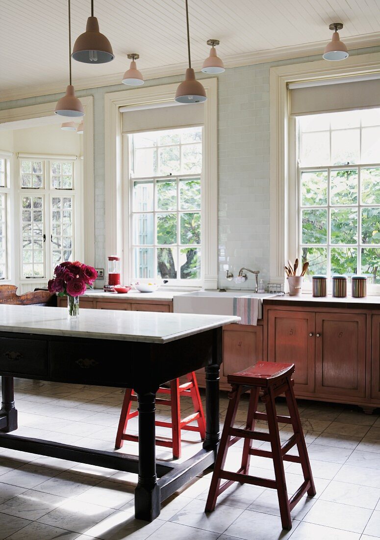 Landhausküche mit Schiebefenstern und Retrolampen, in der Mitte Holzhocker um einen massiven Tisch mit Marmorplatte