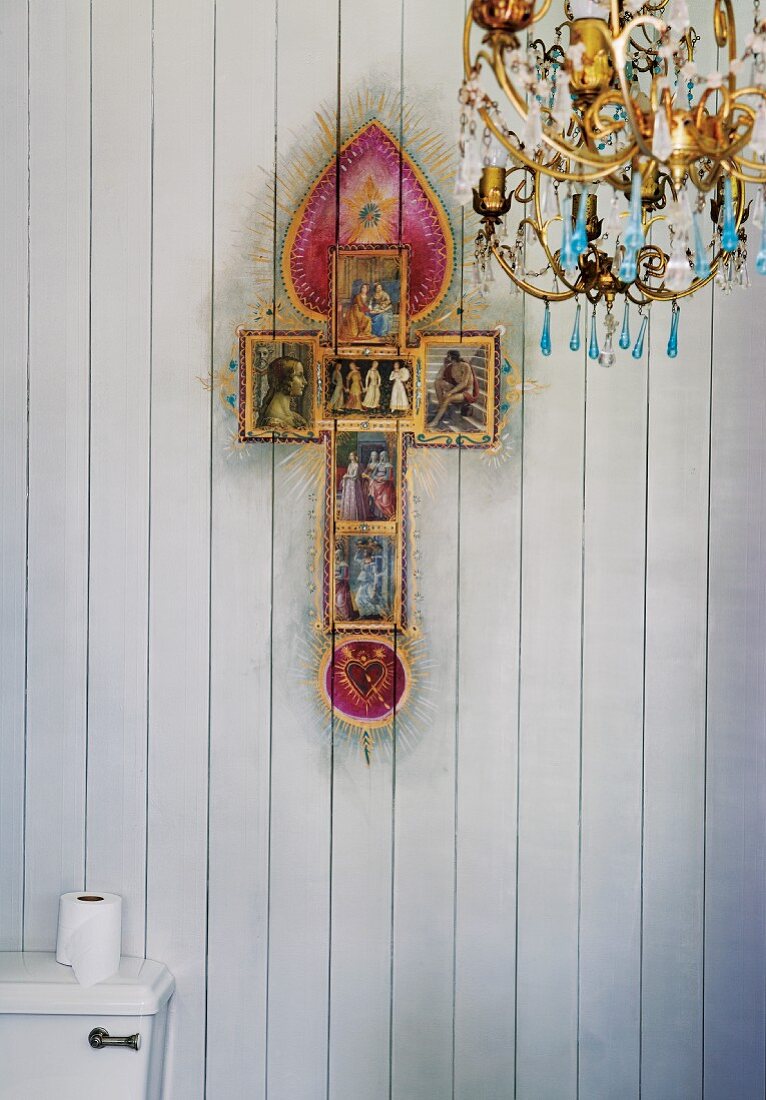 Kitschvariante eines christlichen Kreuzes und Kronleuchter vor holzverkleideter Wand in der Toilette