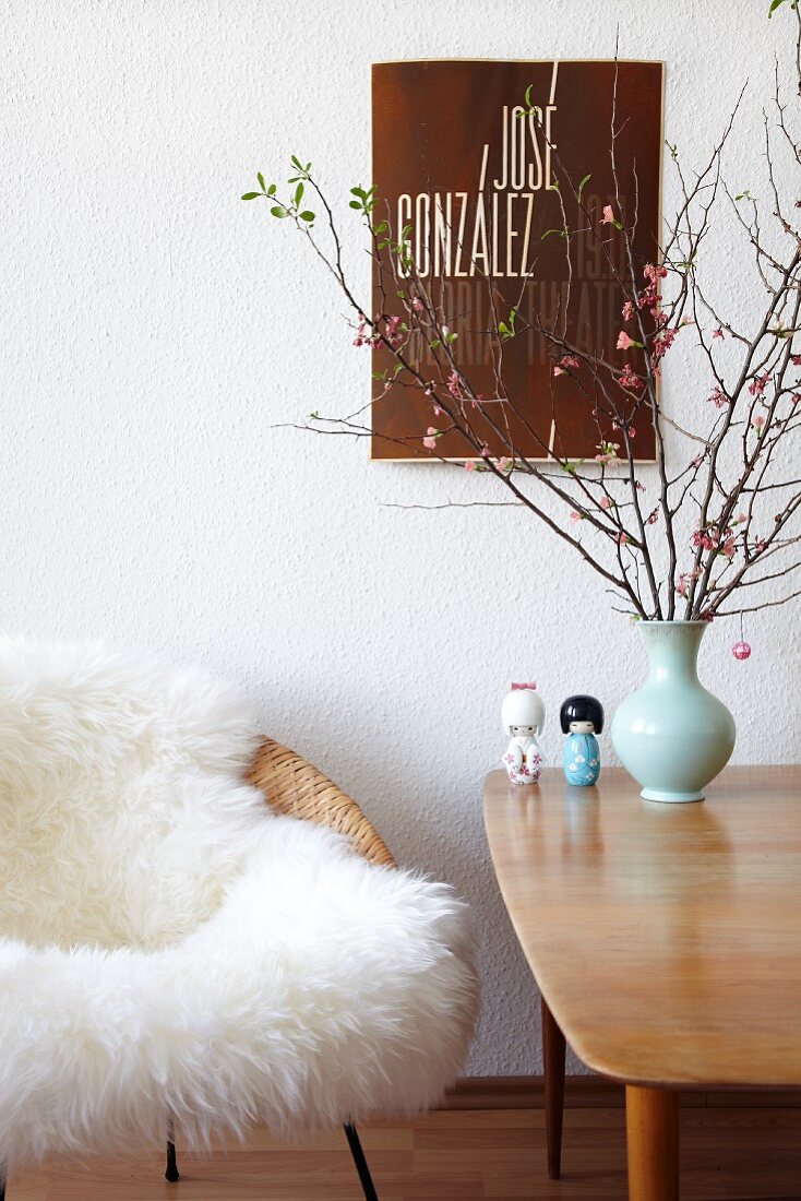 Stuhl mit weißem Tierfell neben Holztisch im Fiftiesstil und Blumenvase vor der Wand mit aufgehängtem Plakat
