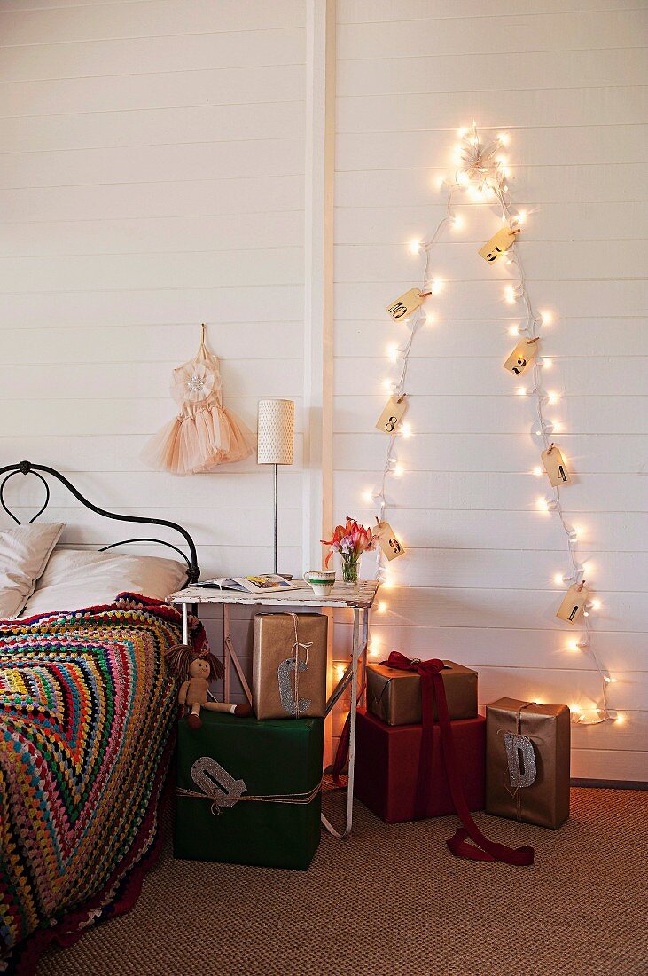 Lichterkette in Weihnachtsbaum-Form mit nummerierten Tütchen an der Wand, davor Geschenkpäckchen