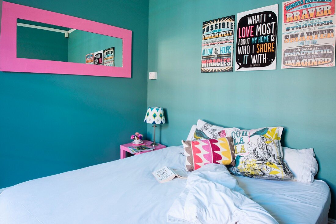 Colourful scatter cushions on bed below pink-framed mirror and artworks with mottoes in English on turquoise wall