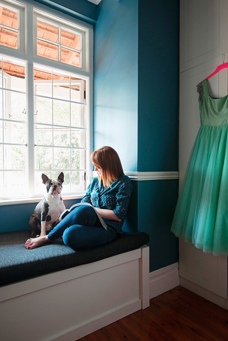 Frau und Hund auf gepolsterter Fensterbank vor blau gestrichener Wand