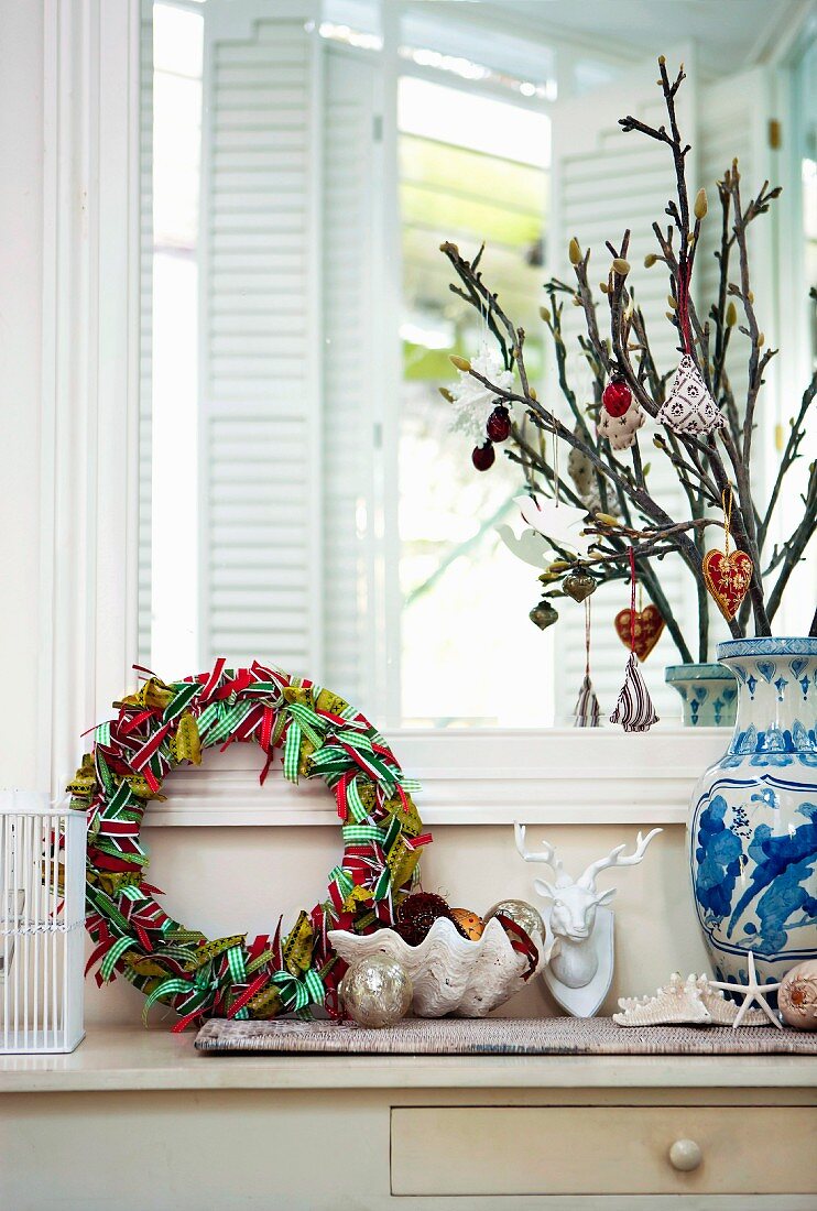 Mit farbigen Bändern umwickelter Adventskranz und Weihnachtsbaumschmuck an Zweigen in einer Keramikvase