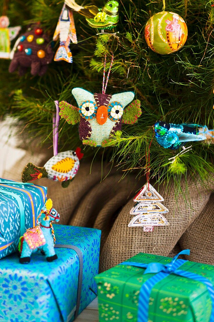 Presents wrapped in blue and green paper below Christmas tree decorated with various, colourful figurines