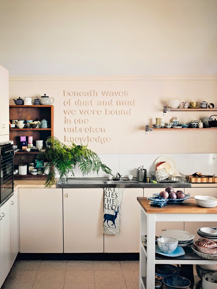 Many items of crockery on wall-mounted shelves and in shelving unit flanking aphorism in relief on kitchen wall