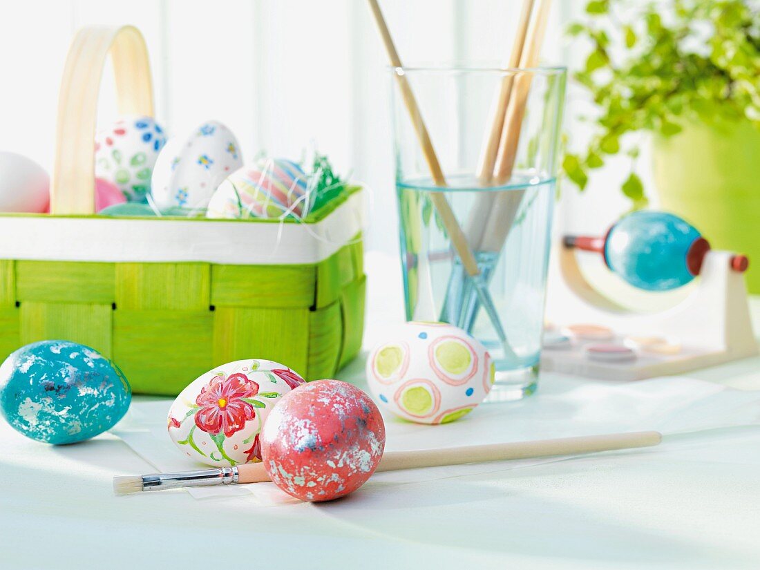 Painted Easter eggs in front of basket of eggs and paintbrushes in glass of water