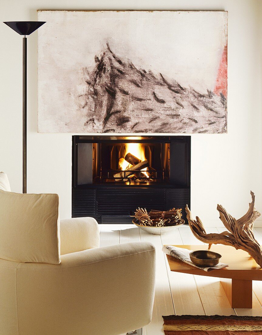 Modernes Gemälde über brennendem Feuer im offenen Kamin; davor ein gemütlicher, weisser Lesesessel