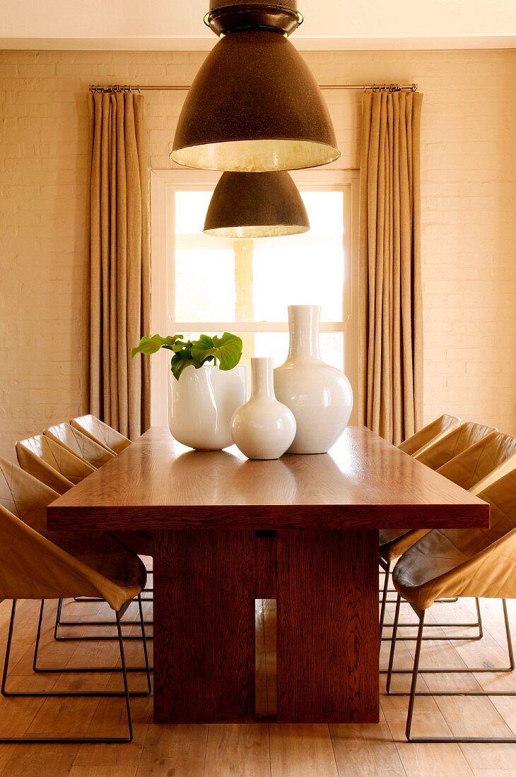 Esstisch aus Massivholz mit weissen Keramikvasen und darüberhängenden, großen Deckenleuchtern vor Esszimmerfenster