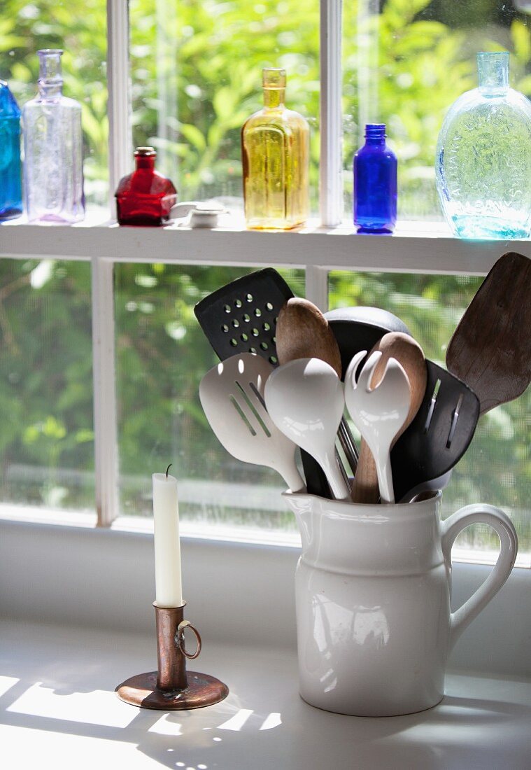 Verschiedene Küchenutensilien in einem Keramikkrug am Küchenfenster, bunte Glasflaschen auf der Fensterbank