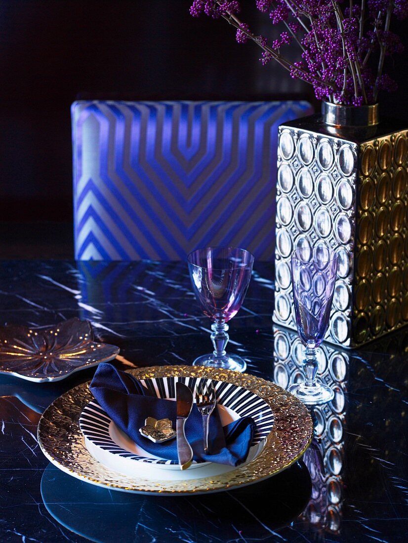 Gedeck mit Goldrandteller, Gläsern, silberglänzender Vase auf Esstisch, Stuhl mit eleganter Polsterlehne