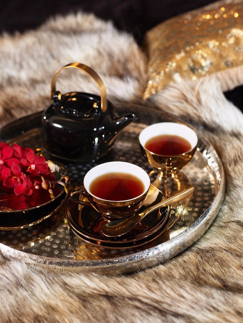 Tablett mit Teetassen, schwarzer Teekanne und roten Orchideenblüten auf Felldecke