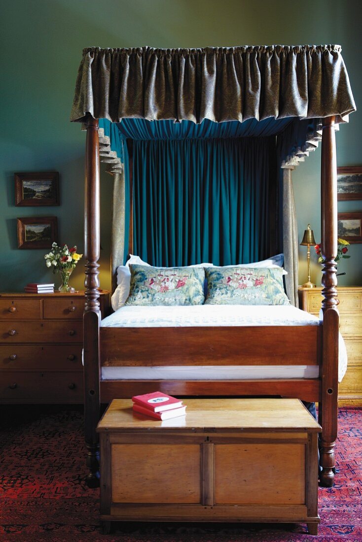 Wooden bed with ruched canopy flanked by bedside cabinets with drawers and with trunk at foot