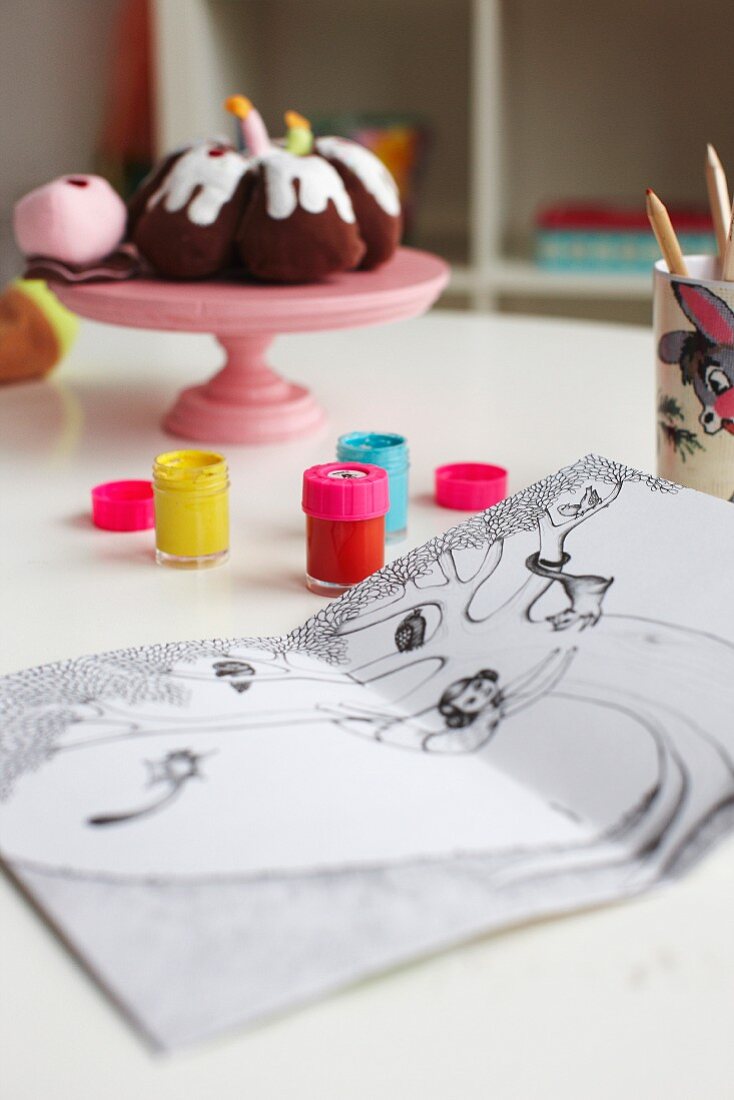 Pots of paints and colouring book on table in child's bedroom; decorative, fabric cakes on plastic cake stand
