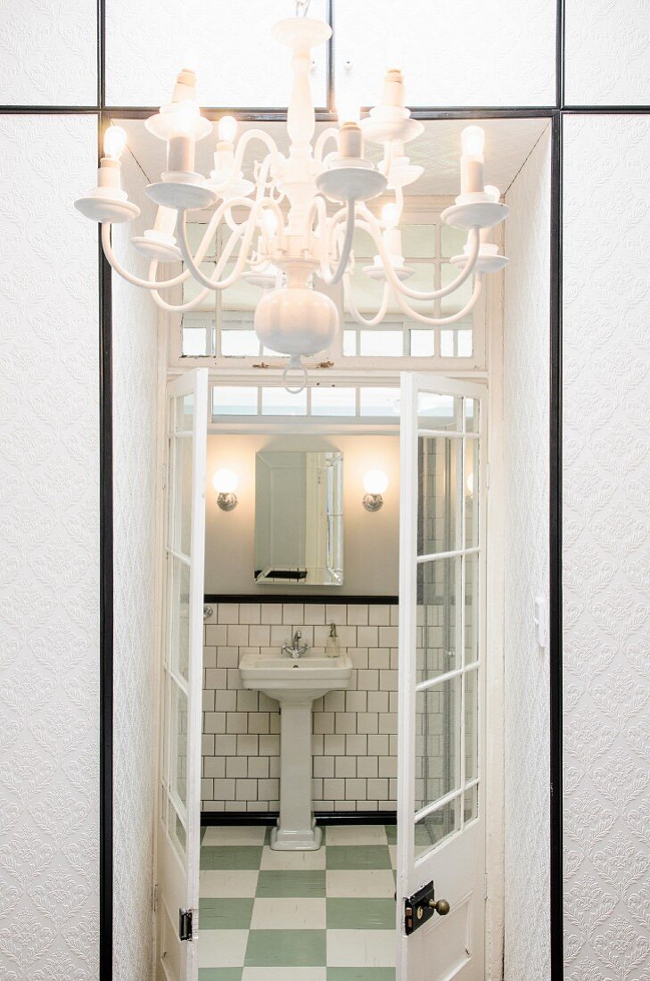 Blick durch geöffnete Sprossenflügeltür ins Badezimmer mit Säulenwaschbecken; weiß tapezierter Einbauschrank und Kronleuchter im Vordergrund