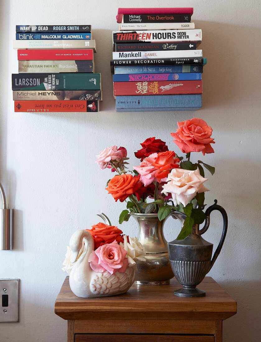 Bouquets of roses in metal jugs and swan-shaped china vase on half-height cabinet below stacked books on minimalist, floating bookshelves