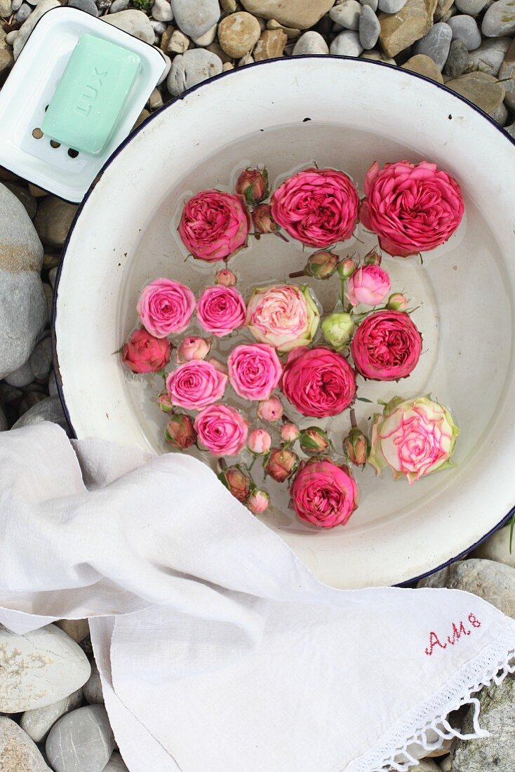 Schwimmende Rosenblüten in alter Emailleschüssel mit weißem Leinenhandtuch und Seifenschale auf einer Kiesfläche arrangiert