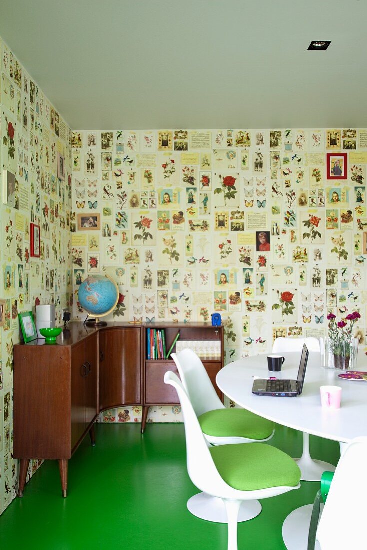 White shell chairs with green seat cushions at white dining table and 50s-style corner sideboard against patterned wallpaper