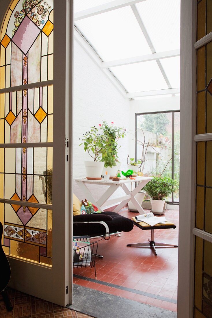 Blick durch offene Flügeltür mit bunter Bleiverglasung in modernen Wintergarten mit Relaxstuhl und Pflanztöpfen