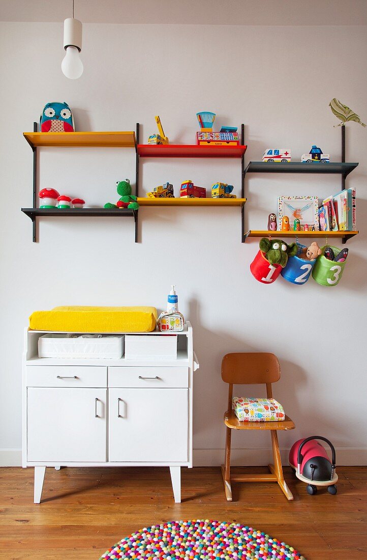 White changing cabinet below colourful shelves of toys on wall and multicoloured felt ball rug
