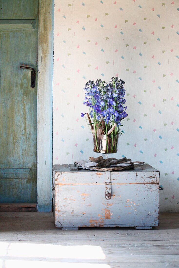 Glass vase of delphiniums and driftwood arranged on vintage trunk against wallpaper with delicate pattern