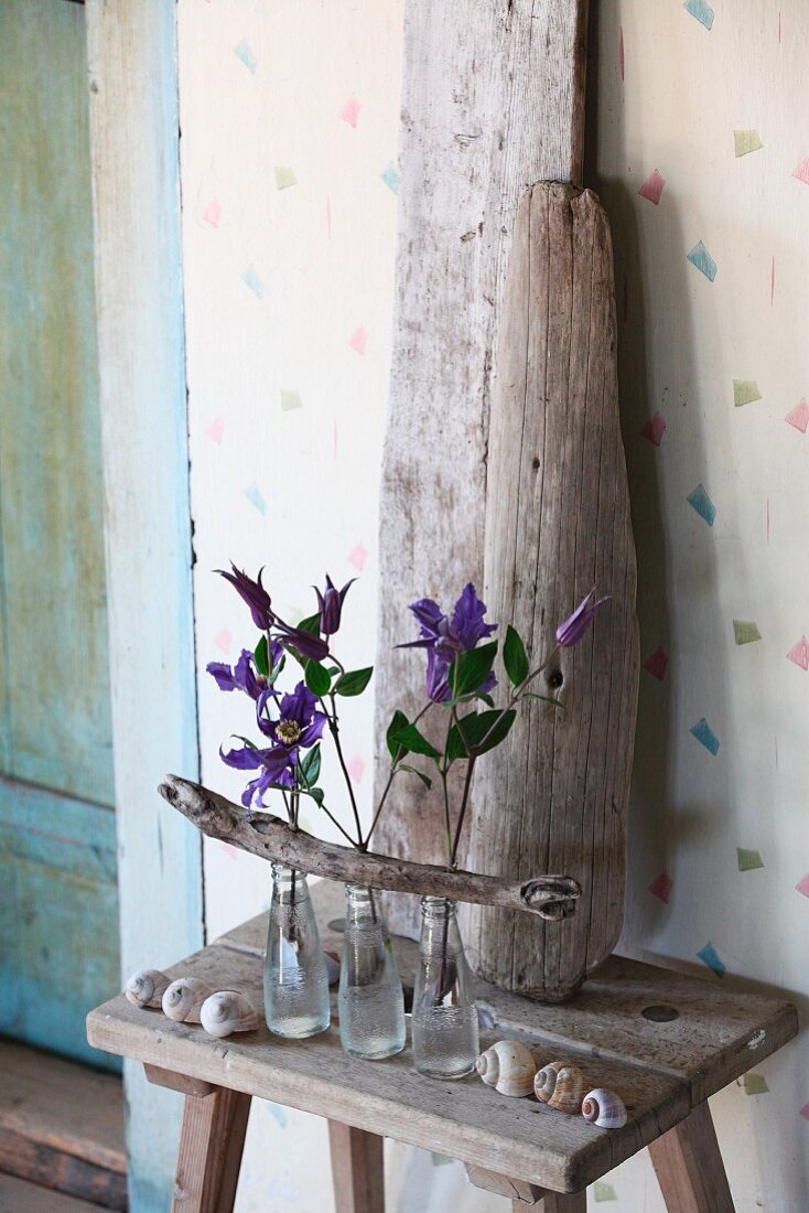 Arrangement of clematis flowers in three glass bottles with driftwood branch in front of upright driftwood pieces on wooden stool in vintage ambiance