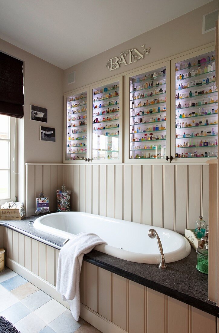 Collection of multicoloured tiny bottles in lit glass-fronted cabinets above fitted bathtub with wood-clad side and stone surround