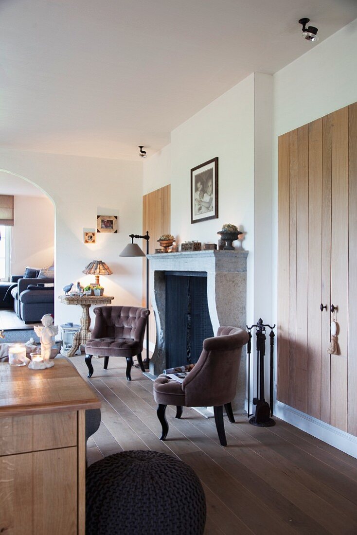 Antique-style armchair in front of open fireplace and fitted cupboards with simple board doors in open-plan interior