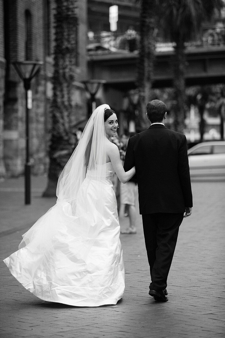 Braut in langem weißem Kleid neben Bräutigam in schwarzem Anzug auf der Straße (s-w-Aufnahme)