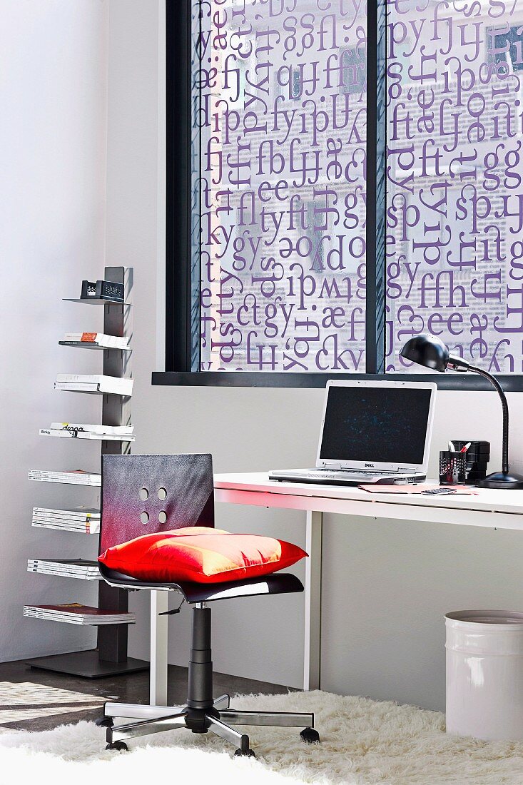 Bürostuhl mit rotem Kissen und minimalistischer Schreibtisch am Fenster, mit Buchstaben bedrucktem Sichtschutz