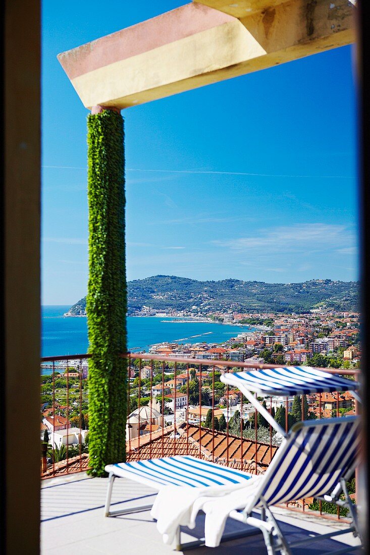 Blauweiss gestreifte Sonnenliege auf Terrasse mit berankter Tragstütze; Blick über eine Bucht an der italienischen Rivieraküste