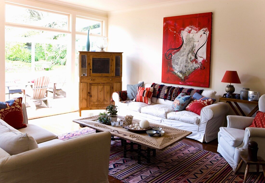 Möbelstücke aus Holz zwischen kissenbestückten Sofas und auffällig rotem Gemälde in Wohnraum mit Blick auf sonnige Terrasse