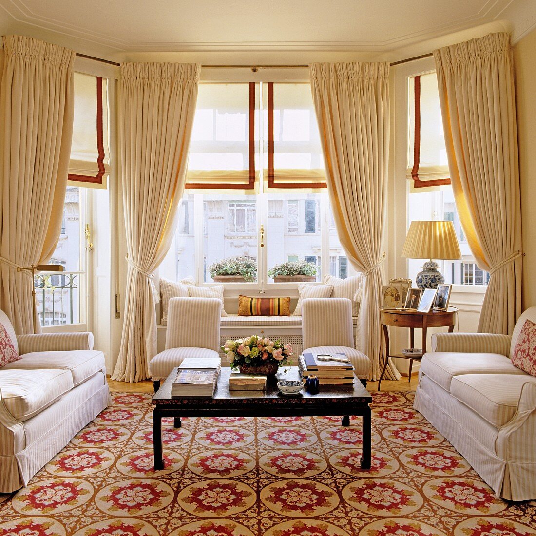 Luxuriously furnished interior with sofa set and coffee table on patterned rug in front of bay window with draped curtains