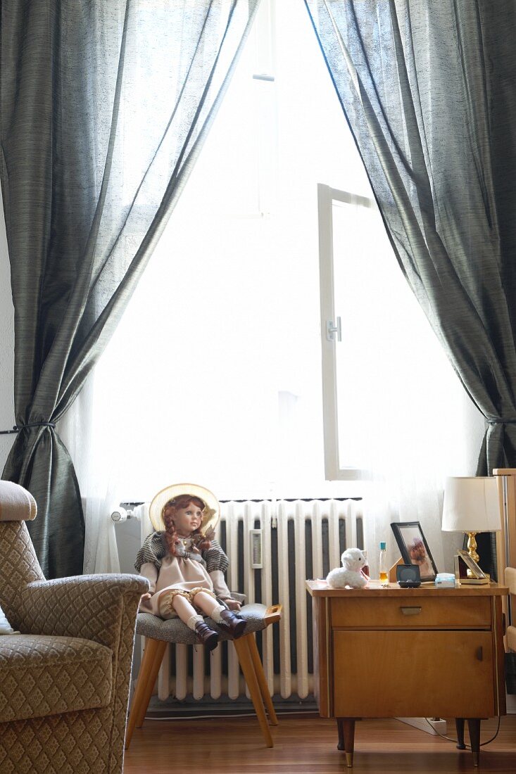 A doll sitting on a stool between an armchair and a night stand in front of a window