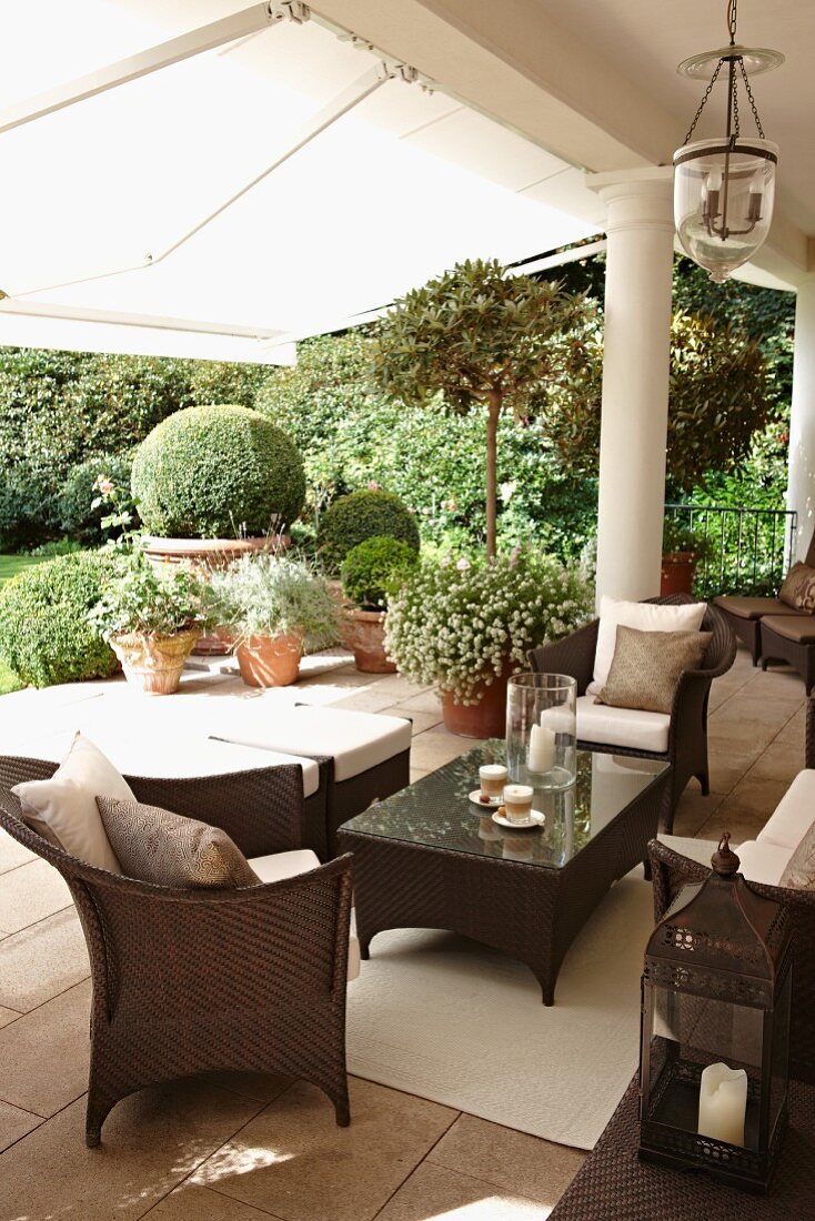 Brown rattan outdoor furniture on a traditional terrace with columns and white awning; view of the garden