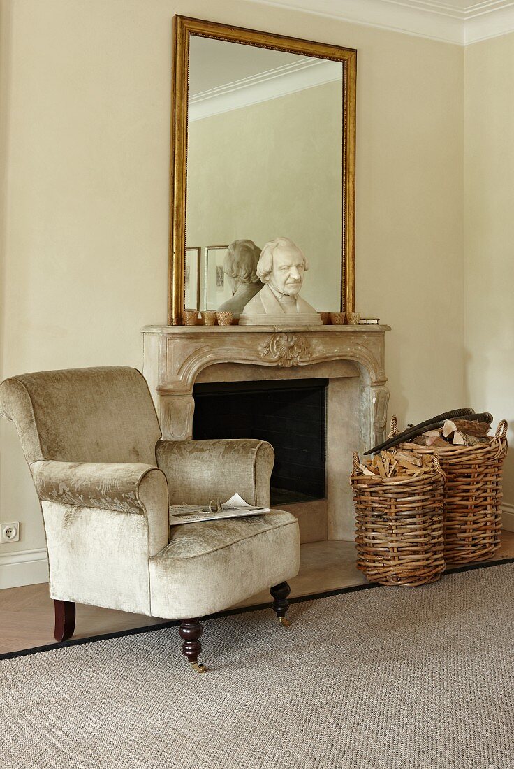 Antique reading armchair covered in gray velvet and wicker basket and firewood in front of an open fireplace with a traditional feel. Large wall mirror on the mantelpiece with a bust in front of it