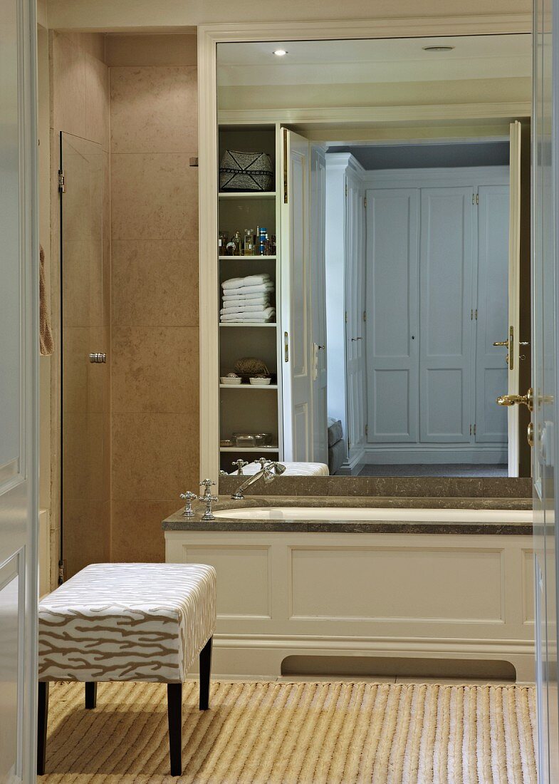 View through and open door of a paneled bathtub in front of a large wall mirror and upholostered stool on a bamboo runner in country home style