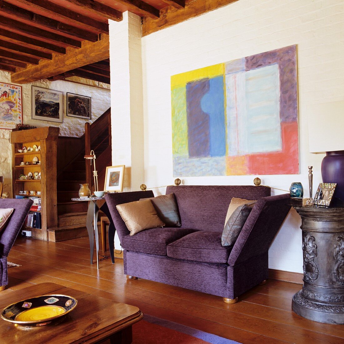 Purple sofa below modern artwork on wall in open-plan interior with wood-beamed ceiling