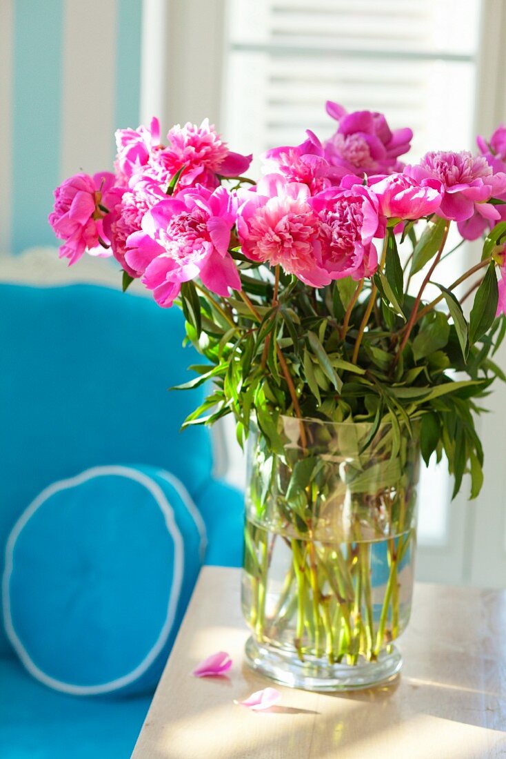 Peonies in a vase on a wooden table in front of an armchair