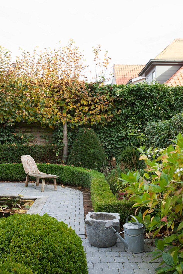 Garden with rustic wooden lounger on paved path and clipped hedges