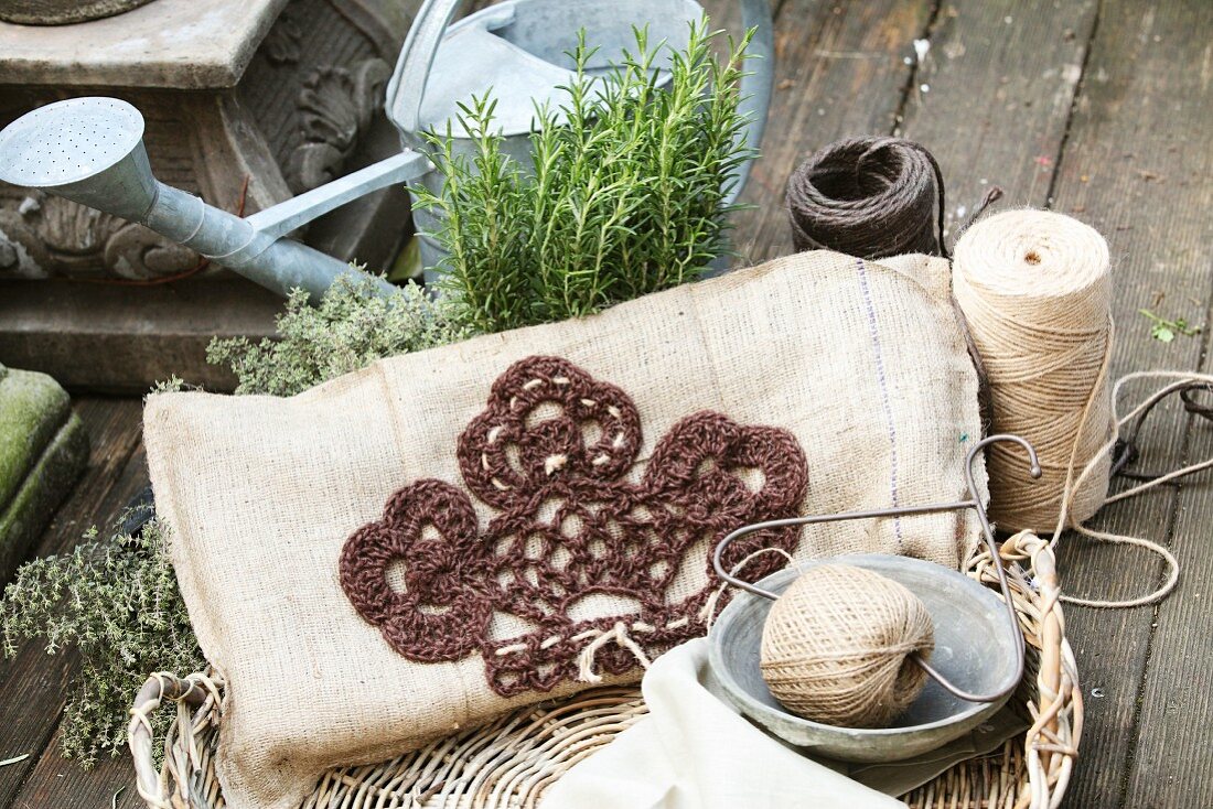 Embroidered cushion next to reels of yarn and herb plants in front of watering can